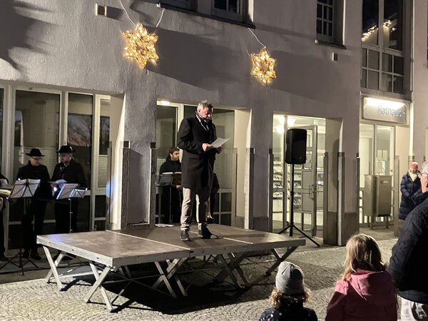 B�rgermeister Mayr steht auf einer kleinen  B�hne und liest Weihnachtsgeschichten. Im Hintergrund steht ein Bl�serensemble, das Rathausgeb�ude ist mit Sternen weihnachtlich dekoriert - im Vordergrund Zuschauer bzw. Zuh�rer